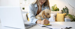 Moça sentada fazendo anotações com uma caneta azul sobre uma mesa, com o notebook ao lado, e segurando um cachorro no colo