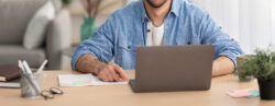 Homem branco com camisa azul utilizando o notebook na sala de sua casa