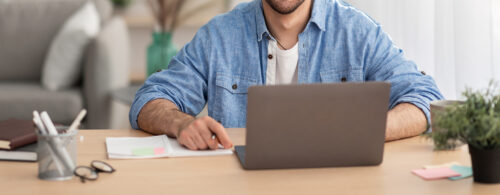 Homem branco com camisa azul utilizando o notebook na sala de sua casa